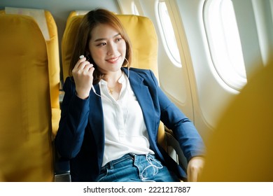 Young Asian Businesswoman Relaxed Listening To Music, Podcast, News, Or Audiobook Using Earphones While Sitting In Airplane Cabin Looking Out The Window. Business And Technology Concept.