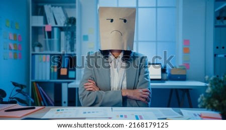 Young Asian businesswoman sit on desk wear draw sad mask paper craft bag feel bad mood with fail work project in office at night. Anonymous lady mad and confuse with overwork, Work mental health.