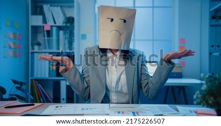 Young Asian businesswoman sit on desk wear draw sad mask paper craft bag feel bad mood with fail work project in office at night. Anonymous lady mad and confuse with overwork, Work mental health.