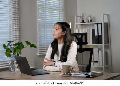 Young asian businesswoman sitting at her desk, arms crossed and gazing thoughtfully away, contemplating business strategies in a modern office environment filled with natural light - Powered by Shutterstock