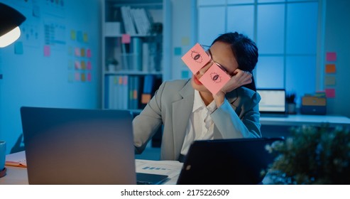 Young Asian businesswoman sitting on desk overworked tired sleep over a laptop at office at night. Exhausted burnout lady with two post-it over her eyes, adhesive notes on face sleeping at workplace.