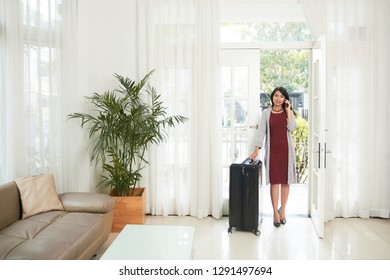 Young Asian businesswoman talking on mobile phone while entering to the modern house with her luggage, she has a business trip - Powered by Shutterstock