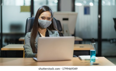 Young Asian Businesswoman Using Laptop Sitting In Modern Office Working With Social Distancing Wearing Mask Alcohol Gel On Desk.