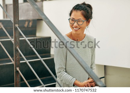 Young Asian businesswoman wearing glasses and smiling while sitting on stairs in an office reading a funny text message on her cellphone