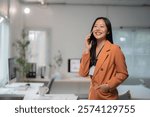 Young Asian businesswoman wearing an orange suit, smiling and talking on her smartphone while standing confidently in a modern office, hand casually tucked in her pocket