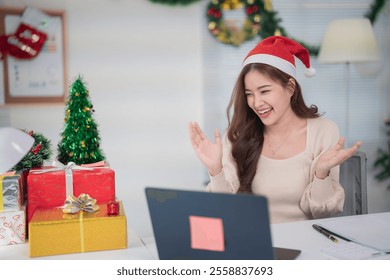 Young asian businesswoman wearing a santa hat video calling colleagues while celebrating christmas at her desk, surrounded by festive decorations and gift boxes - Powered by Shutterstock