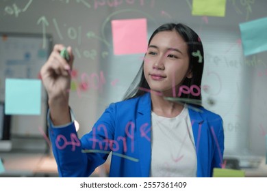 Young asian businesswoman writing calculations and using sticky notes on a transparent glass board in the office - Powered by Shutterstock