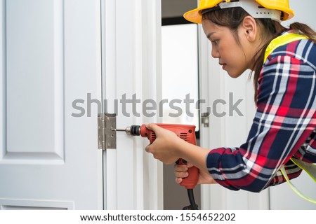 Young Asian carpenter woman, technician or worker using power drill bit installing screwed a door hinges or hinges on wooden door. House, Renovation or Drilling Equipment Construction service concept