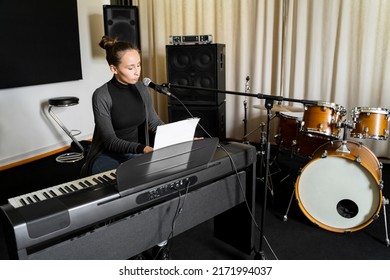 Young Asian Or Caucasian Brunette Woman Singing Song On Mic And Playing Digital Keyboard Electronic Piano Alone Near Drums In Music Studio. Getting Ready For Exam In Conservatory Or Music School