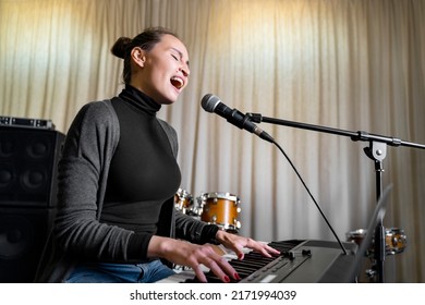 Young Asian or Caucasian brunette woman having fun singing song on mic emotionally loud with pleasure and playing digital keyboard electronic piano in music studio or school, event or concert stage