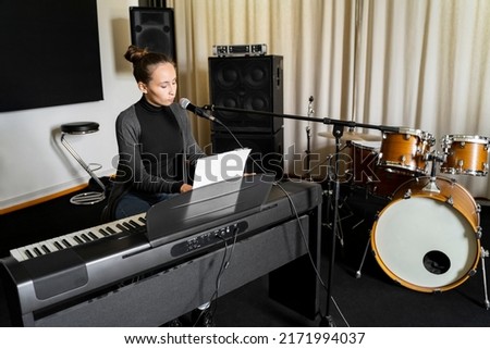 Young Asian or Caucasian brunette woman singing song on mic and playing digital keyboard electronic piano alone near drums in music studio. Getting ready for exam in conservatory or music school Foto d'archivio © 