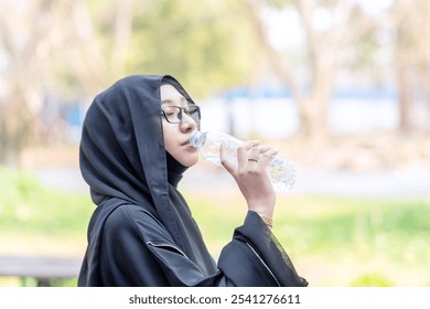 Young Asian cheerful Muslim woman in hijab traditional clothes hold a water bottle and drink in publics park on blurred background. - Powered by Shutterstock