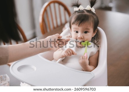 Young Asian child eats while seated in a high chair, lovingly assisted by a parent. A scene representing care and nourishment during early childhood development.