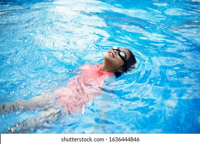 Young Asian Child With Peach Swimwear Floating In Pool.Swimming Lesson. Girl With Goggles In Blue Water.Smiling Kid Having Fun During Vacation. Backstroke.