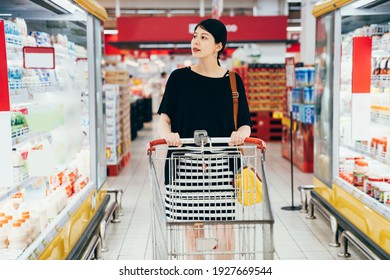 Young asian chinese charming pregnant woman pushing shopping cart with grocery products shelves on side. beautiful future motherhood buying food with trolley in store. elegant parent in supermarket