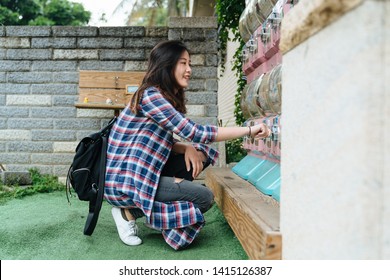 Young Asian Chinese Girl Traveler Kneeling Down On Grass Lawn Outdoor Playing Japanese Capsule Toy Machine In Japan. Beautiful Teenager Female With Backpack Having Fun In Farmer Market Insert Coin