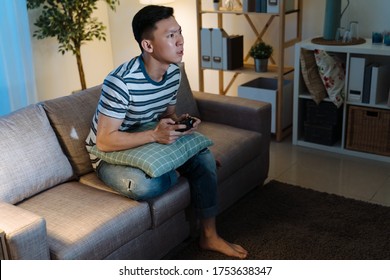 Young Asian Chinese Man Addicted To Video Gaming At Dark Home. Concentrated Handsome Guy In Casual Wear Stay Up Late House Living Room. Male Sitting On Sofa Holding Joystick Remote In Evening.