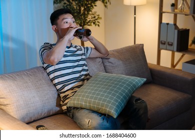 Young Asian Chinese Man Watching Television Drinking Bottle Of Beer In Late Evening Time. Handsome Guy Enjoy Alcohol Alone In Sofa At Night In Apartment. Single Male Stay Up Late Relax On Couch