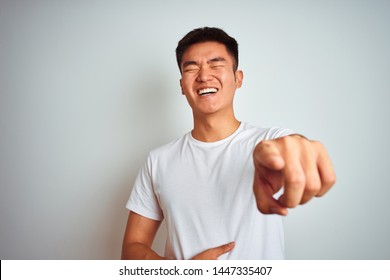 Young asian chinese man wearing t-shirt standing over isolated white background laughing at you, pointing finger to the camera with hand over body, shame expression