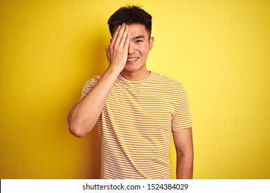 Young asian chinese man wearing t-shirt standing over isolated yellow background covering one eye with hand, confident smile on face and surprise emotion.