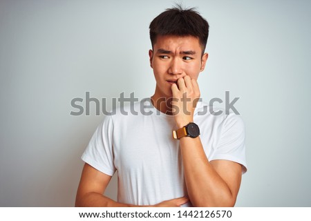 Young asian chinese man wearing t-shirt standing over isolated white background looking stressed and nervous with hands on mouth biting nails. Anxiety problem.