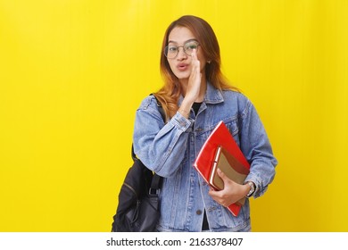 Young asian college student standing while holding books and whispering. Isolated on yellow