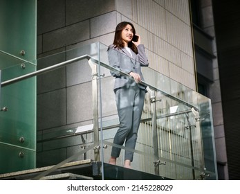 Young Asian Corporate Business Woman Standing On Top Of Stairs Making A Call Using Cellphone In Modern Office Building