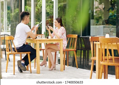 Young Asian couple checking social media and texting friends when having breaksfast in outdoor cafe