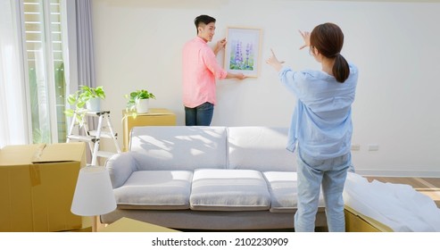 young asian couple decorate home - man hanging photo with frame on wall and woman helping him in living room