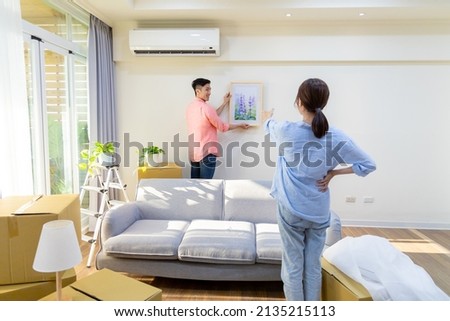young asian couple decorate home - man hanging photo with frame on wall and woman helping him in living room