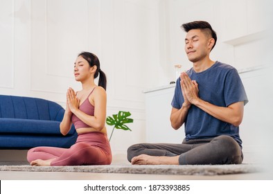 Young Asian Couple Doing Meditation Yoga In Living Room At Home Closing Eyes And Breathing Slowly, Concentrate Practicing Yoga Lotus Position. Relax Breathe Easy Seat Pose Home Healthy Lifestyle.