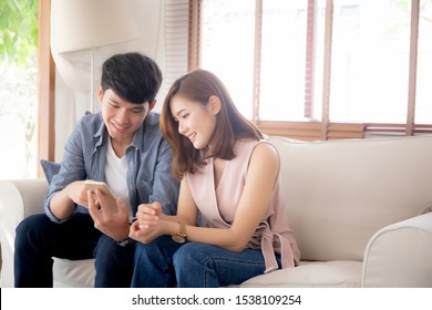 Young Asian Couple Or Friend Looking Smart Mobile Phone With Enjoy At Living Room, Happy Family Relax And Fun Media Entertainment On Smartphone On Sofa Together At Home, Lifestyle Concept.