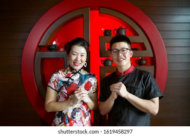 Young Asian couple greeting. Chinese New Year celebration in Asia. Husband and wife wearing traditional China costume. Lady wearing qipao. Waiter and waitress in Oriental restaurant.Chef at work.