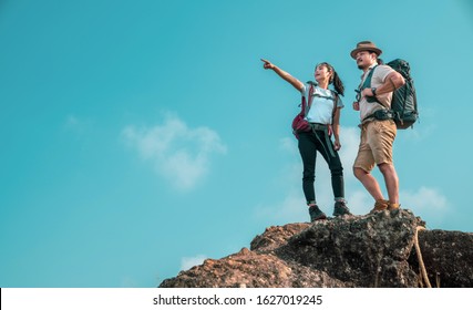 Young Asian Couple Hikers Climbing Up On The Peak Of Mountain. Two People Hikers On Top Of The Mountain Enjoying Valley View. Tourists With Backpacks. Climbing ,Helps And Team Work Concept