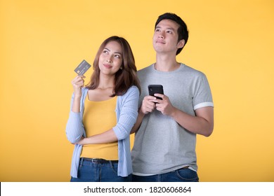 Young Asian couple holding credit card and smartphone while looking up isolated on yellow background