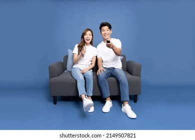 Young Asian Couple Holding Remote Tv And Watching Television On A Sofa Isolated On Blue Background
