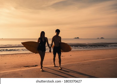 Young Asian Couple Holding Surfboard On The Beach At Morning