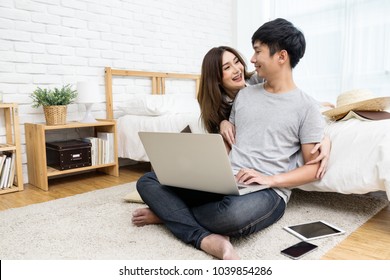 Young asian couple looking together using computer laptop internet shopping online or searching data honeymoon trip travel at bedroom in house.Happy lover hug each other and smiling on bed at  home