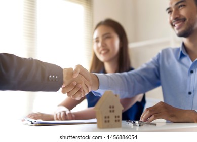 Young Asian couple making contract with house sale agency. Man shaking hand with female agent sitting opposite and his wife sitting next to him with smile looking at house broker. Real estate concept.