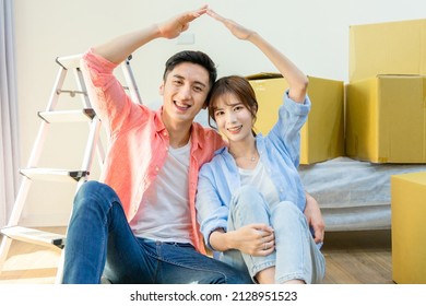 Young Asian Couple Moving Into New Home - They Sitting On Wooden Floor Are Using Their Arm To Form A Home Sign