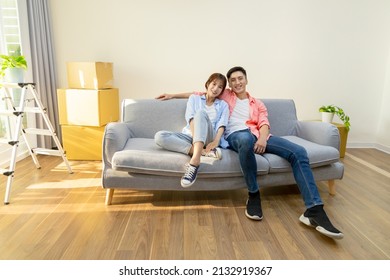 Young Asian Couple Moving Into New Home - They Are Sitting On Sofa And Smiling At You