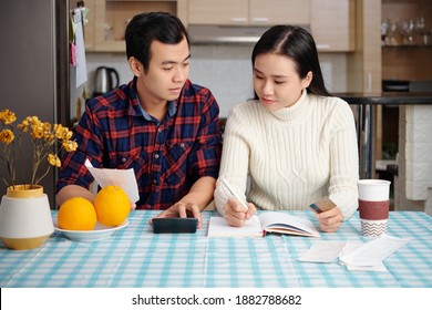 Young Asian Couple Reviewing Bills, Planning Family Budget And Calculating Finances At Kitchen Table With Bills, Calculator And Planner