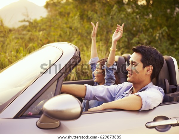 Young Asian Couple Riding In A Convertible Sport Car Enjoying The Cool Breeze At Sunset 