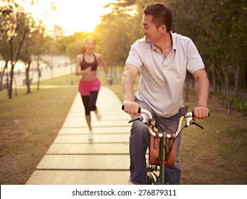 Young Asian Couple Running, Riding Bike Outdoors In Park At Sunset, Fitness, Sport And Exercise, Healthy Life And Lifestyle Concept.