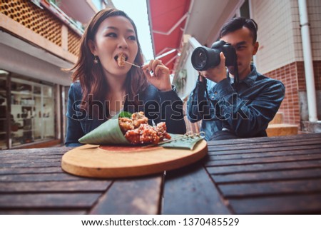 Young asian couple sitting in chinese cafe outside. At front on wooden table is traditional asian food - bamboo leaf wrap. Man is taken a photo of a woman. Woman is posing for him, enjoying asian food
