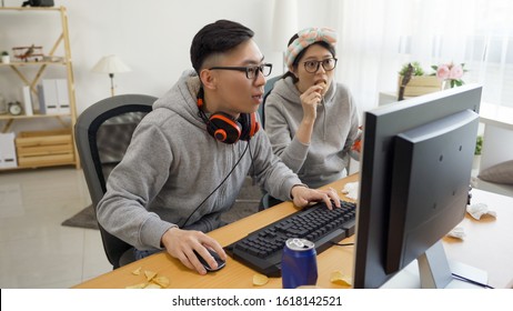 Young Asian Couple Sitting In Messy Home Indoorsy Homebody Relationship. College Student Girlfriend And Boyfriend Playing Online Computer Game Together On Pc Screen Eating Chips Snack On Summer Break