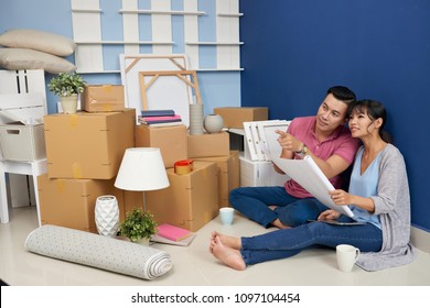 Young Asian Couple Sitting On The Floor In New Flat With Cardboard Boxes, Holding Blueprint Of Room And Discussing Furniture Arrangement