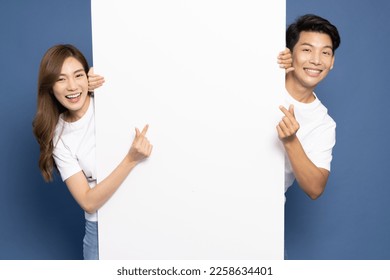 Young Asian couple is standing behind the white blank banner or empty copy space advertisement board and showing mini heart sign isolated on blue background, Looking at camera - Powered by Shutterstock