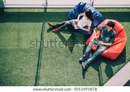 Young Asian couple using laptop notebook and digital tablet together in modern public park, sit on bean bag, top view with copy space on grass. Information technology gadget or casual business concept