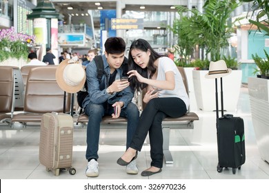 Young Asian Couple Using Smart Phone While Sitting In The Airport Terminal Waiting For Boarding. 